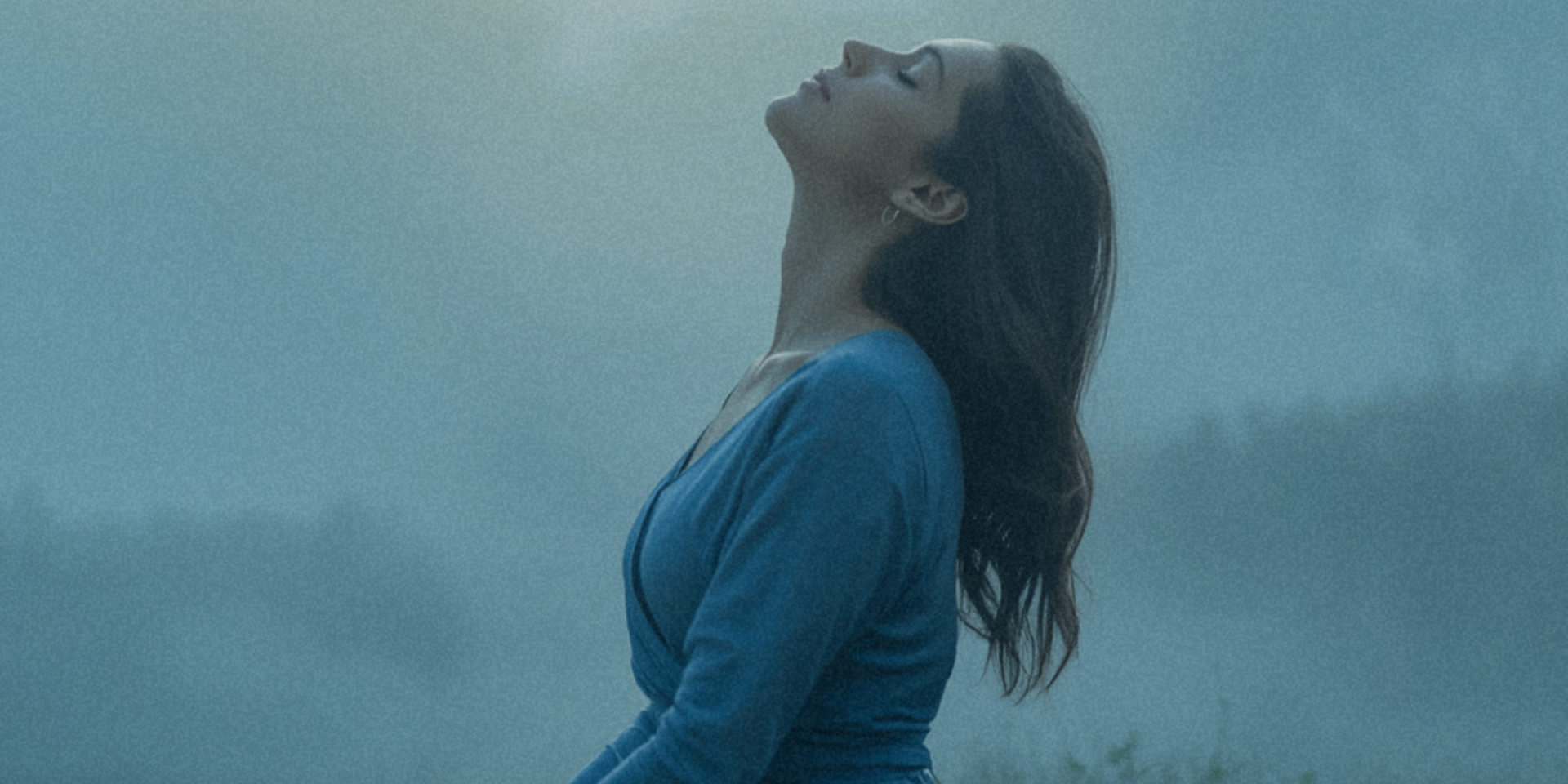 Woman standing with eyes closed and head tilted back, breathing deeply in a soft blue mist